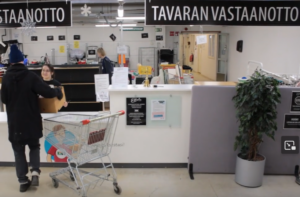 Elävä kauppa Kuopion tavaran vastaanotto pisteellä kolme henkilö. Lahjoittaja tuo tavaraa vastaanottoon työntekijälle. Lajittelija häärii taustalla.